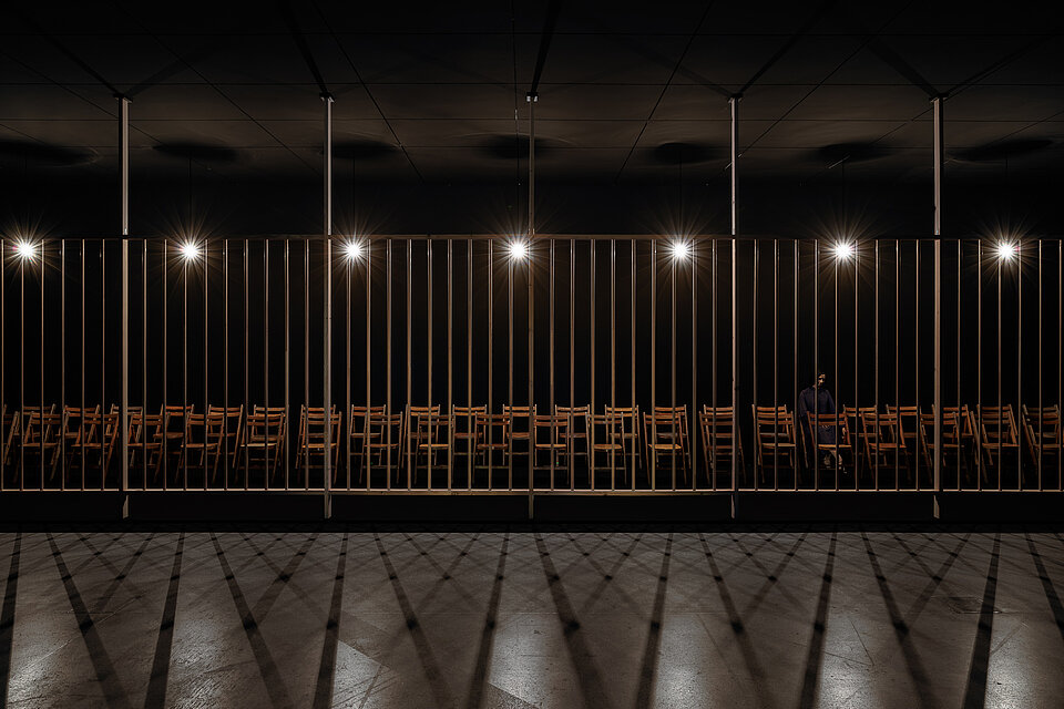 
            
                Dunkler Raum mit einer langen Reihe aufgestellter Holzstühle hinter einer hohen Gitterstruktur
            
        