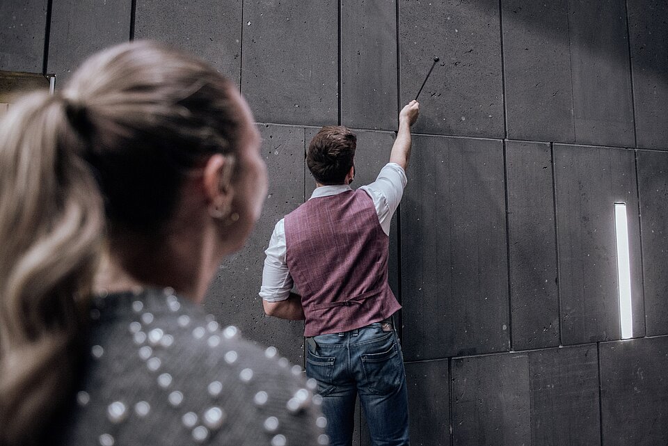 
            
                eine Frau von hinten mit langen Haaren, ein Mann zeigt auf eine dunkle Steinwand
            
        