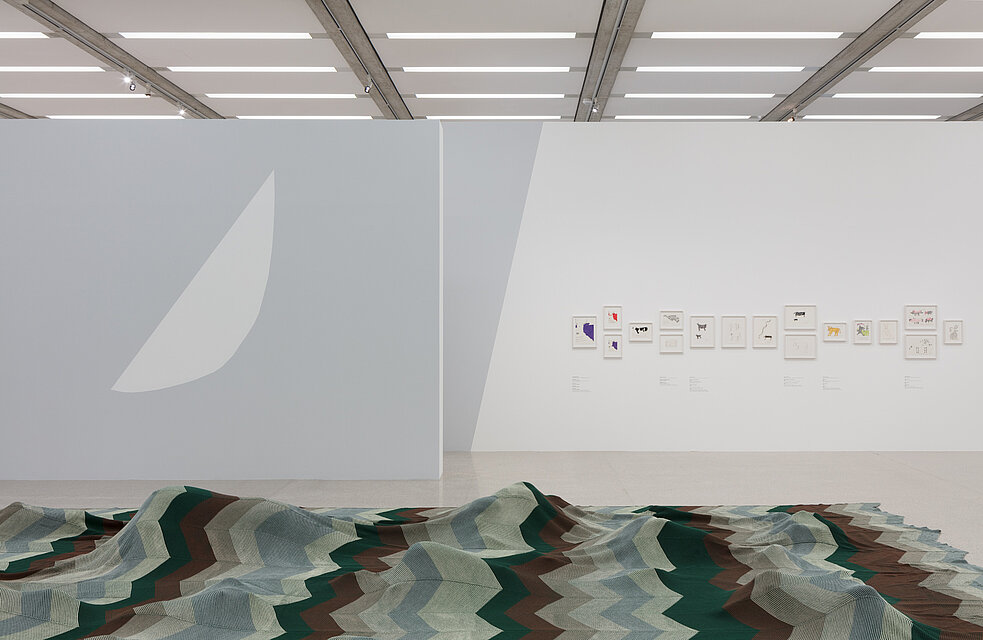 A large-format piece of fabric with a brown, green and blue wave pattern. Lying in the centre of the floor. On the exhibition wall behind it hang twelve smaller pictures with abstractly painted animal motifs.