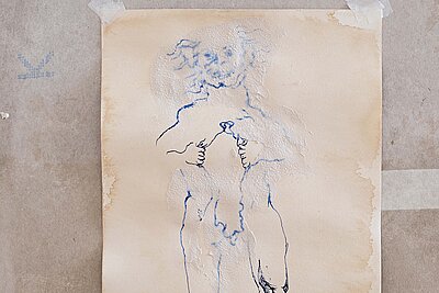 Aufgeklebtes Blatt mit verblasster blauer Linienzeichnung einer stehenden menschlichen Figur auf strukturierter, teils fleckiger Wandfläche. / Taped sheet showing a faded blue line drawing of a standing human figure on a textured, partially stained wall surface.
