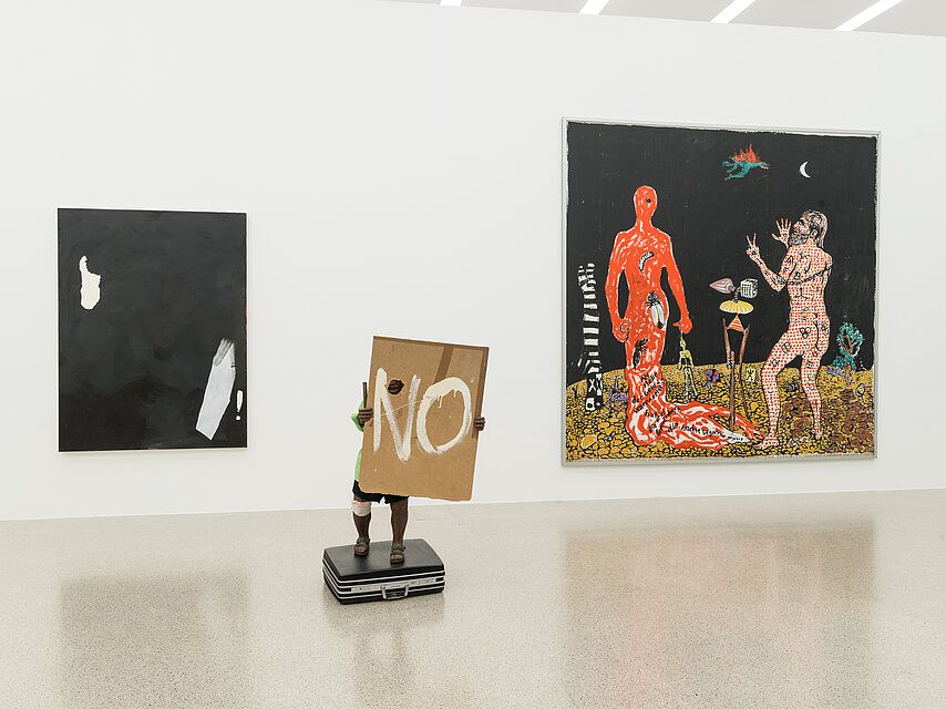 On the left is a black picture on the wall, in the middle a small sculpture holding up a sign with "NO", on the right a dark picture with two colourful figures on it