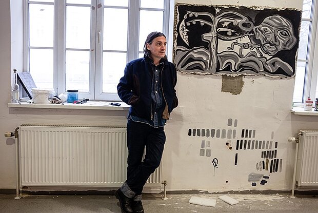 
            
                Künstler Tobias Pils in seinem Atelier, rechts hängt ein Kunstwerk, links befindet sich ein Fenster
            
        