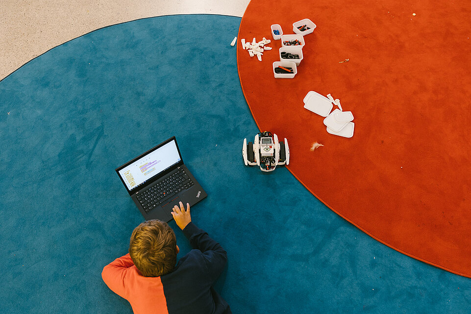 Kind liegt am Boden und schaut in einen Laptop, daneben ein Roboter