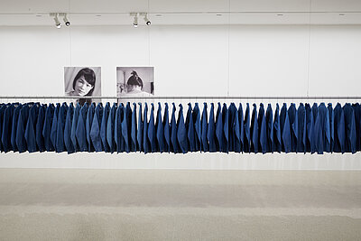 The picture is filled by a clothes rail with countless denim shirts. In the background 2 black and white portraits of women