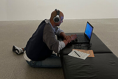 junger Mann sitzt am Boden, trägt Kopfhörer und arbeitet an einem Laptop, der auf einer Bank steht
