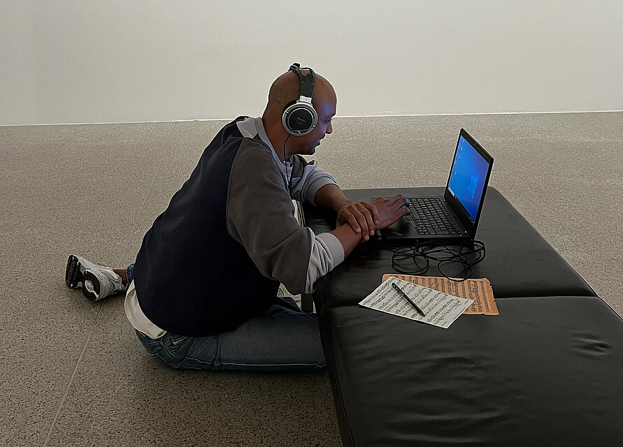 junger Mann sitzt am Boden, trägt Kopfhörer und arbeitet an einem Laptop, der auf einer Bank steht