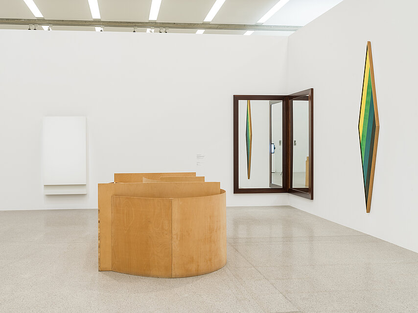 Ausstellungsraum mit einem hölzernen, runden Objekt in der Mitte, rechts dahiner ein Spiegel in der Ecke, rechts an der Wand ein grünes Objekt