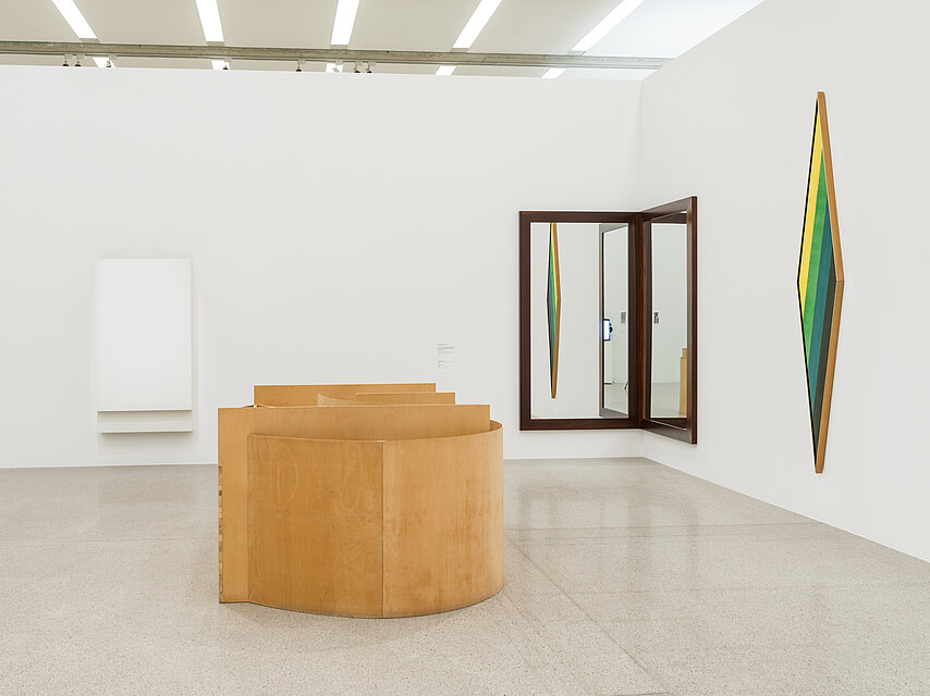  Exhibition room with a wooden, round object in the centre, a mirror in the corner to the right behind it, a green object on the wall to the right