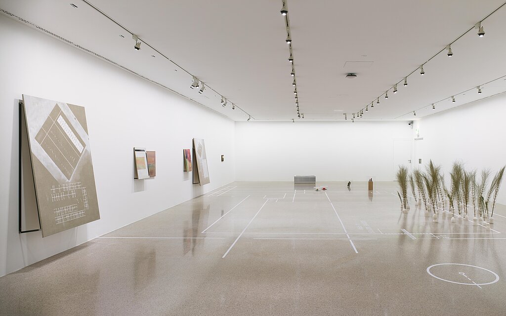 On the right-hand side of the large exhibition room are feather grasses in small white vases. Works of art hang on the left wall. 