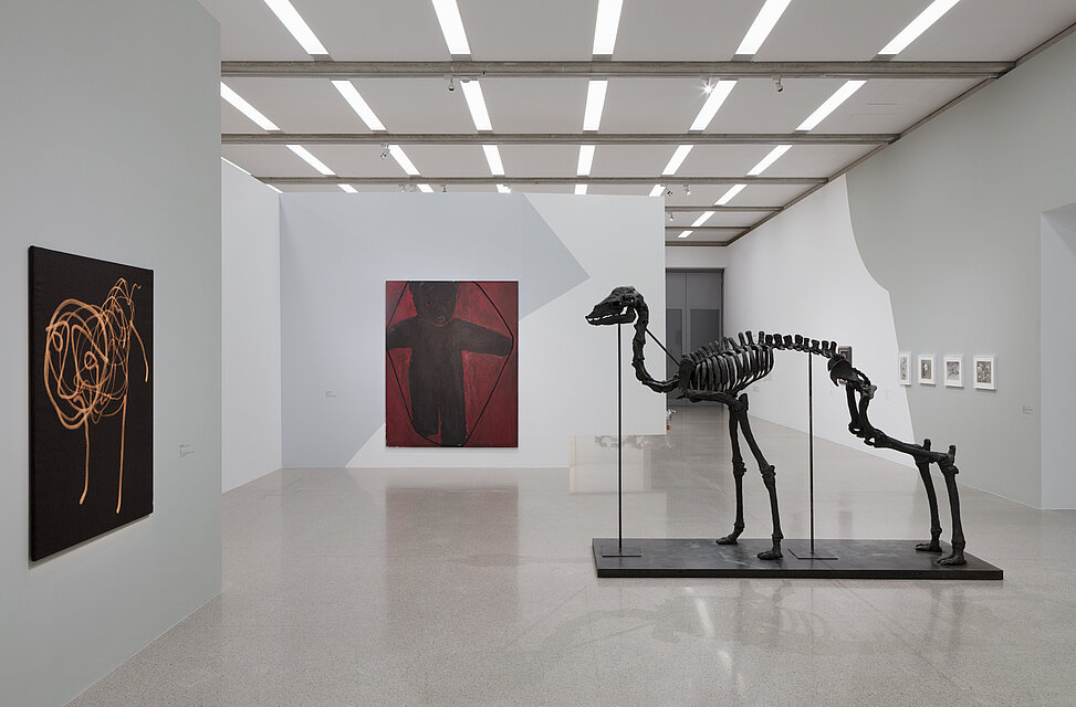 A black painting with the yellow outline of an animal hangs on the left-hand wall of the exhibition; in the background is a red and black painting depicting a bear. On the right side of the room is an artificial skeleton of a camel. 