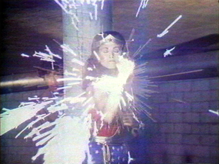 Videostill einer Person vor gemauerter Kulisse, im Moment eines grellen Licht- und Funkenausbruchs, der aus einem gehaltenen Objekt hervorgeht. / Video still of a person in front of a brick-like background at the moment of a bright burst of light and sparks emerging from an object being held.