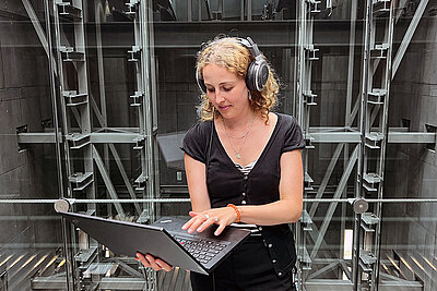 junge Frau steht im mumok Treppenhaus, trägt Kopfhörer und arbeitet an einem Laptop