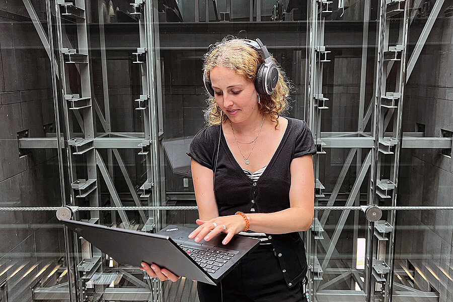 junge Frau steht im mumok Treppenhaus, trägt Kopfhörer und arbeitet an einem Laptop