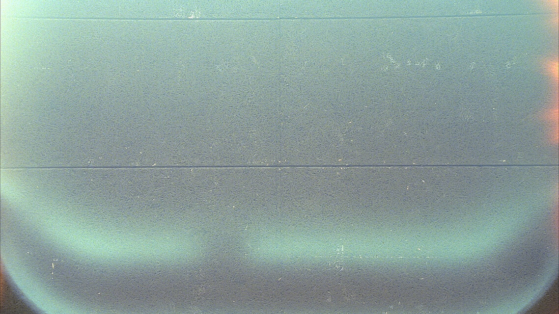 A grainy surface in pale green to bluish tones with visible film marks and horizontal lines. The lower area features a softly curved, brighter zone.