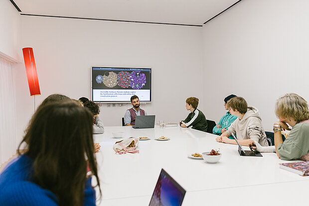 
            
                Kinder und Jugendliche sitzen um einen großen weißen Tisch versammelt, im Hintergrund ein Bildschirm zum Workshop und ein Kunstvermittler mit Laptop
            
        