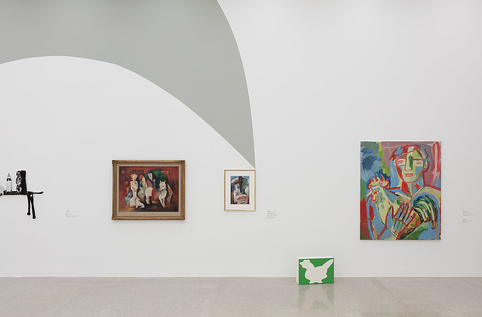 Drei abstrakte, bunte Gemälde mit Tierdarstellungen hängen auf der Wand. Davor steht eine kleine weiß-grüne Box mit einer Hahnabbildung auf dem Boden. 
