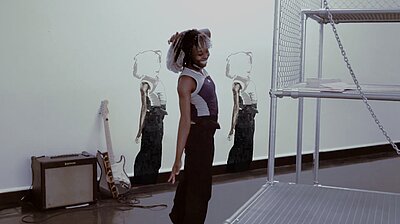 A black dancer stands in a room, her right hand stretched downwards. In front of her is part of a scaffold. In the background, two schematic drawings can be seen on the wall, which look like the dancer and assume the same pose.