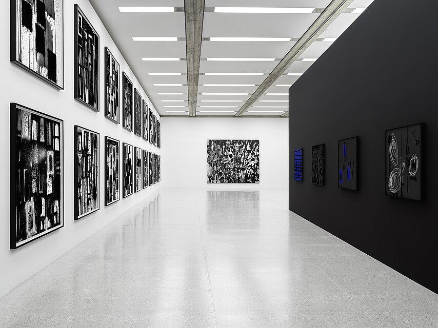 View of an exhibition by Adam Pendleton, light grey floor, left and centre white walls with black and white abstract artworks, right a black wall with dark artworks, occasional blue tones