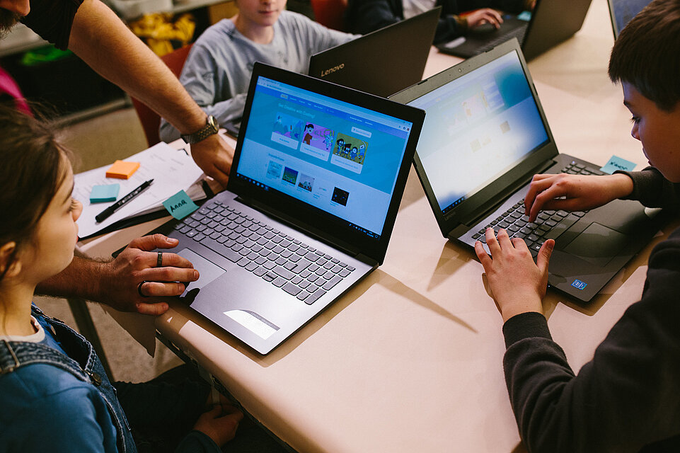 a white table with several open laptops, children sitting around it and working on the laptops