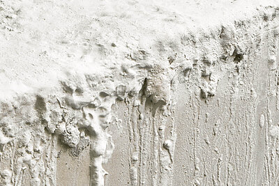 Nahaufnahme einer strukturierten weißen Wandoberfläche mit dicken Farbschichten, Tropfen und unregelmäßigen Erhebungen. / Close-up of a textured white wall surface with thick paint layers, drips, and irregular protrusions.