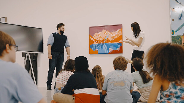 
            
                eine Gruppe Kinder sitz am Boden, wir sehen sie nur von hinten. Zwei Erwachsene erklären den Kindern ein Kunstwerk
            
        