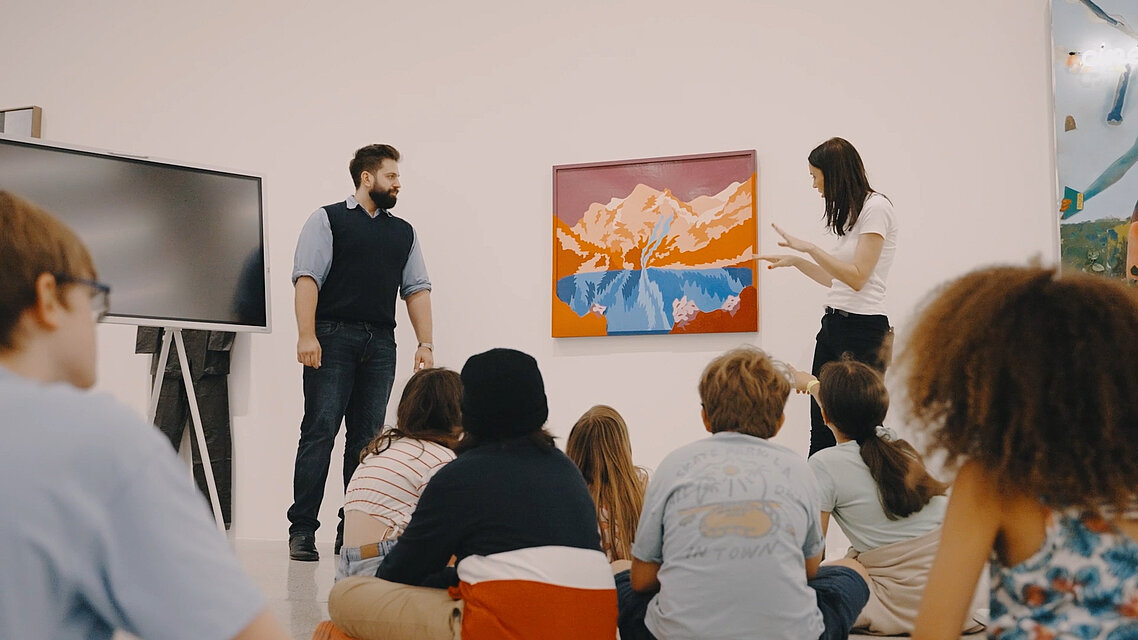 eine Gruppe Kinder sitz am Boden, wir sehen sie nur von hinten. Zwei Erwachsene erklären den Kindern ein Kunstwerk