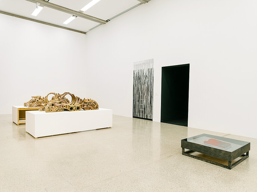 a bright exhibition space, in the background a wide white base, on it a kind of wooden, flat installation, on the right a passage leads into a black room