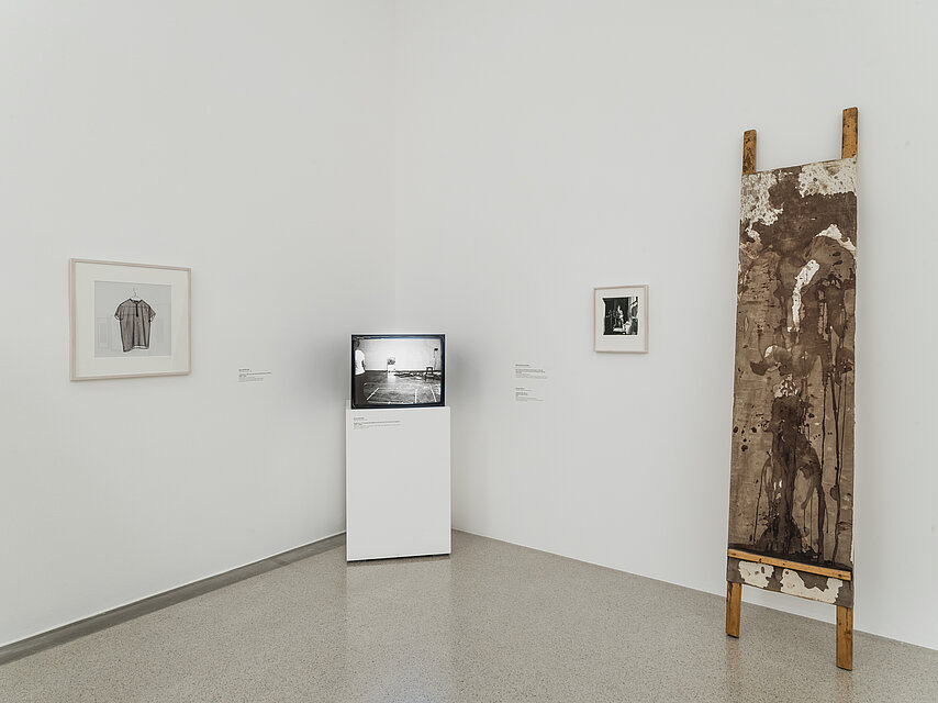 Fernseher auf einem weißen Sockel in einer Ecke, links und rechts davon je ein kleines Bild an der Wand, ganz rechts ein hölzernes, längliches Objekt, das an der Wand lehnt