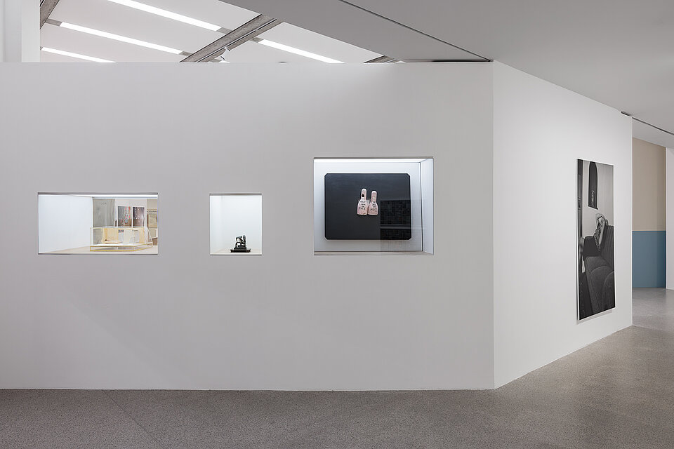 
            
                Weißer Ausstellungsraum mit drei Vitrinen, die unterschiedliche Objekte präsentieren
            
        