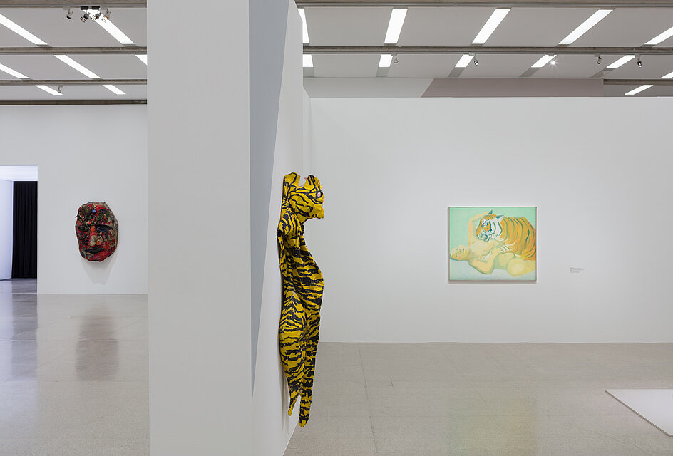 A three-dimensional wall sculpture in the shape of a tiger. Behind it on the right is a painting of a nude and a tiger. In the background on the left is an abstract work of art in the shape of a red face. 