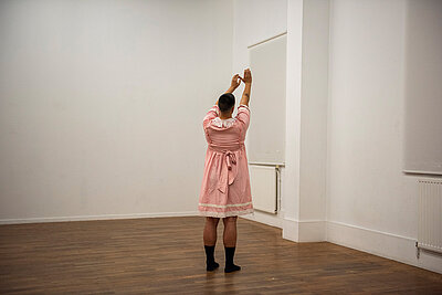 Person in einem leeren Raum mit Holzboden und weißen Wänden, von hinten aufgenommen, trägt ein rosafarbenes Kleid und hebt beide Arme über den Kopf. / Person in an empty room with wooden flooring and white walls, photographed from behind, wearing a pink dress and raising both arms above the head.