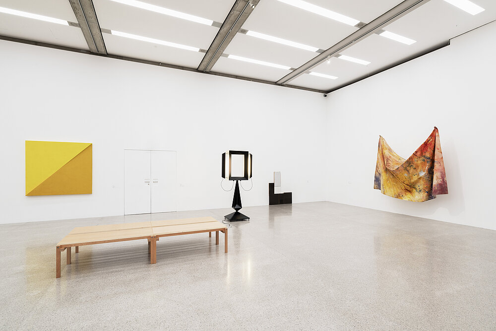 A bright exhibition space in which several works of art can be seen: on the right, a yellow and red cloth hangs loosely from the white wall, suspended at two ends. Two black sculptures in the back corner, a yellow abstract painting on the left. A wooden bench can be seen in the foreground.