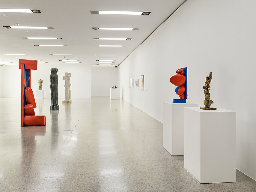 View of an exhibition room with a light-coloured stone floor and white walls, on the right are two small sculptures on white bases on the wall, on the left are elongated sculptures on the floor, the one in front is red, the ones behind are black and white