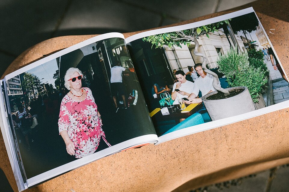 Aufgeschlagenes Fotobuch auf einer Stein- oder Betonoberfläche mit einer Doppelseite, die Straßenszenen zeigt, links eine gehende Person im Vordergrund, rechts mehrere sitzende Personen an einem Tisch im Außenraum. / An open photo book placed on a stone or concrete surface, showing a double-page spread with street scenes, a walking person in the foreground on the left and several seated people at an outdoor table on the right.