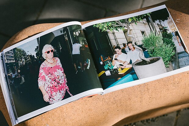 
            
                Aufgeschlagenes Fotobuch auf einer Stein- oder Betonoberfläche mit einer Doppelseite, die Straßenszenen zeigt, links eine gehende Person im Vordergrund, rechts mehrere sitzende Personen an einem Tisch im Außenraum. / An open photo book placed on a stone or concrete surface, showing a double-page spread with street scenes, a walking person in the foreground on the left and several seated people at an outdoor table on the right.
            
        