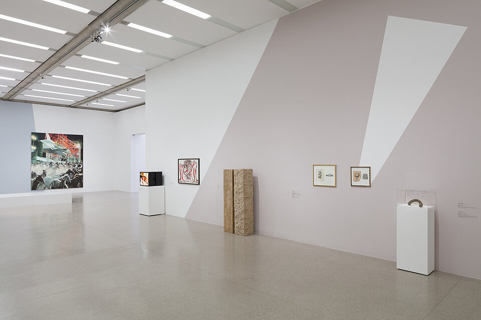 Two works of art on a white base. In between, three pictures and a brown sculpture. A large-format picture in the background.