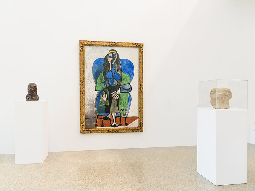 View of an exhibition room with white walls, in the centre an abstract painting of a woman in green and blue dresses by Picasso, on the left and right white bases with small sculptures