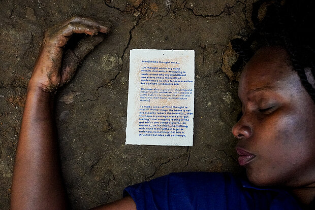 
            
                 Rehema Chachage, What the soil gave us: Eine Person liegt auf Erdboden, neben ihrem Kopf links befindet sich ein Blatt Papier mit Text
            
        