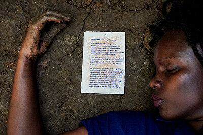  Rehema Chachage, What the soil gave us: Eine Person liegt auf Erdboden, neben ihrem Kopf links befindet sich ein Blatt Papier mit Text
