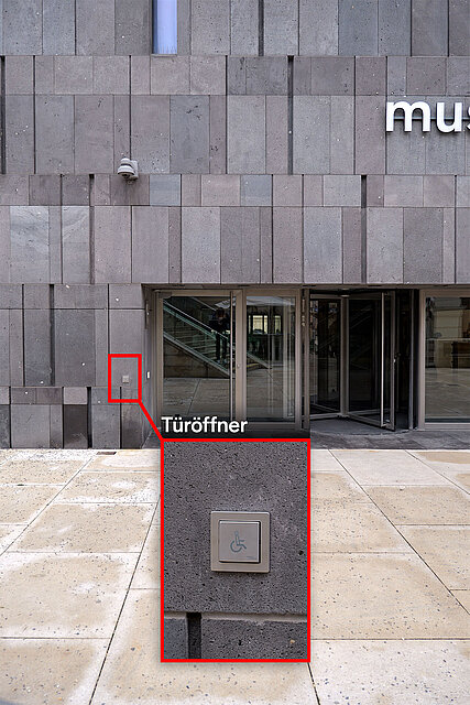 Fassadenansicht eines Gebäudeeingangs mit automatischen Türen, hervorgehoben ist ein barrierefreier Türöffner‑Taster an einem Betonblock vor dem Eingang. / Facade view of a building entrance with automatic doors, highlighting an accessible door opener button on a concrete block in front.