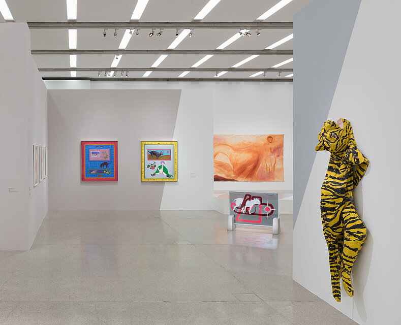 Three colourful paintings in the background. An abstract work of art in front of it. On the right in the foreground, a sculptural depiction of a tiger on the wall.