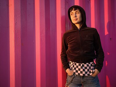A portrait photo of the artist: she is standing in front of a pink wall. She is wearing a black hooded vest with the hood on her head. She is also wearing short denim pants.