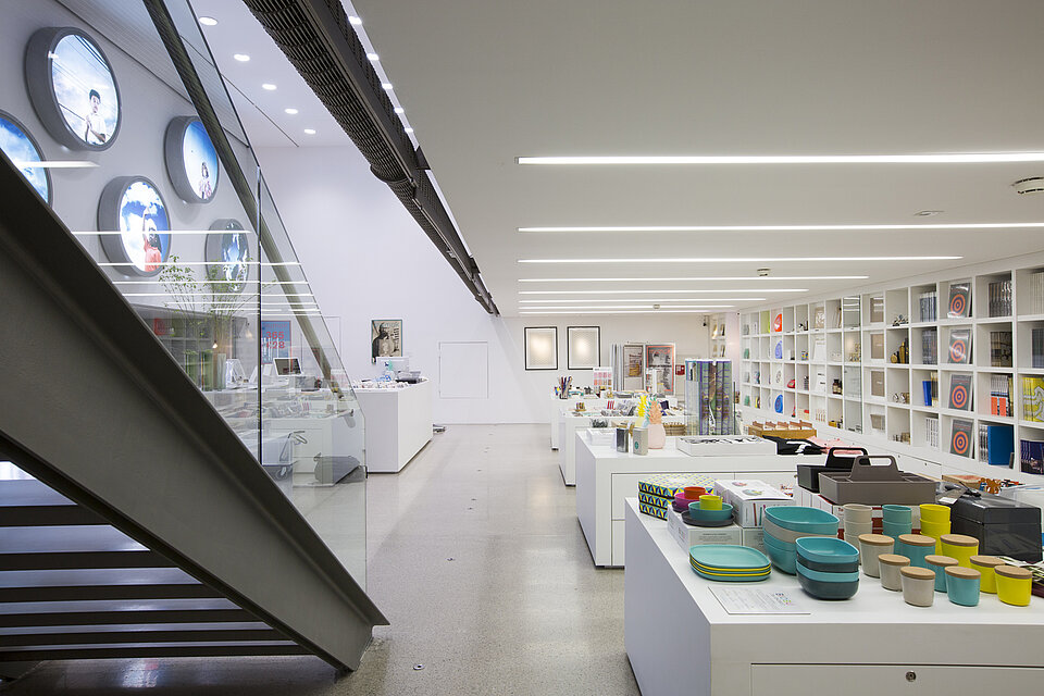 Heller Museumsshop mit weißen Regalen und Tischen, auf denen Bücher, Haushaltswaren und Designobjekte ausgestellt sind, links eine Treppe und runde Wanddisplays mit Porträts. / Bright museum shop with white shelving and tables displaying books, household items, and design objects, with a staircase on the left and circular wall displays with portraits.