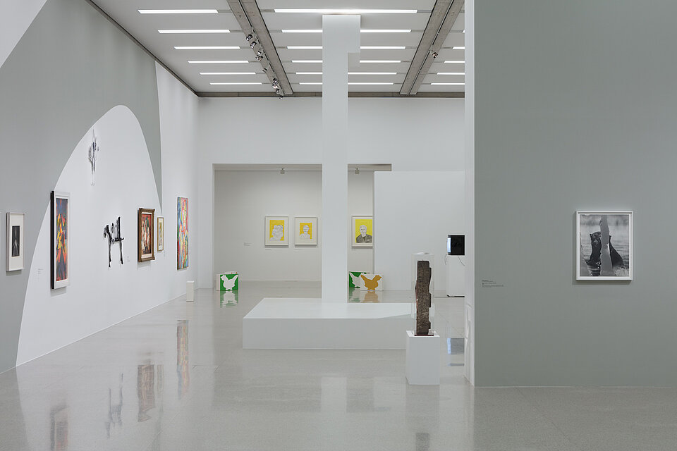 White-grey exhibition walls with paintings in different formats and colours. White pedestals with abstract sculptures are placed in the centre of the room.