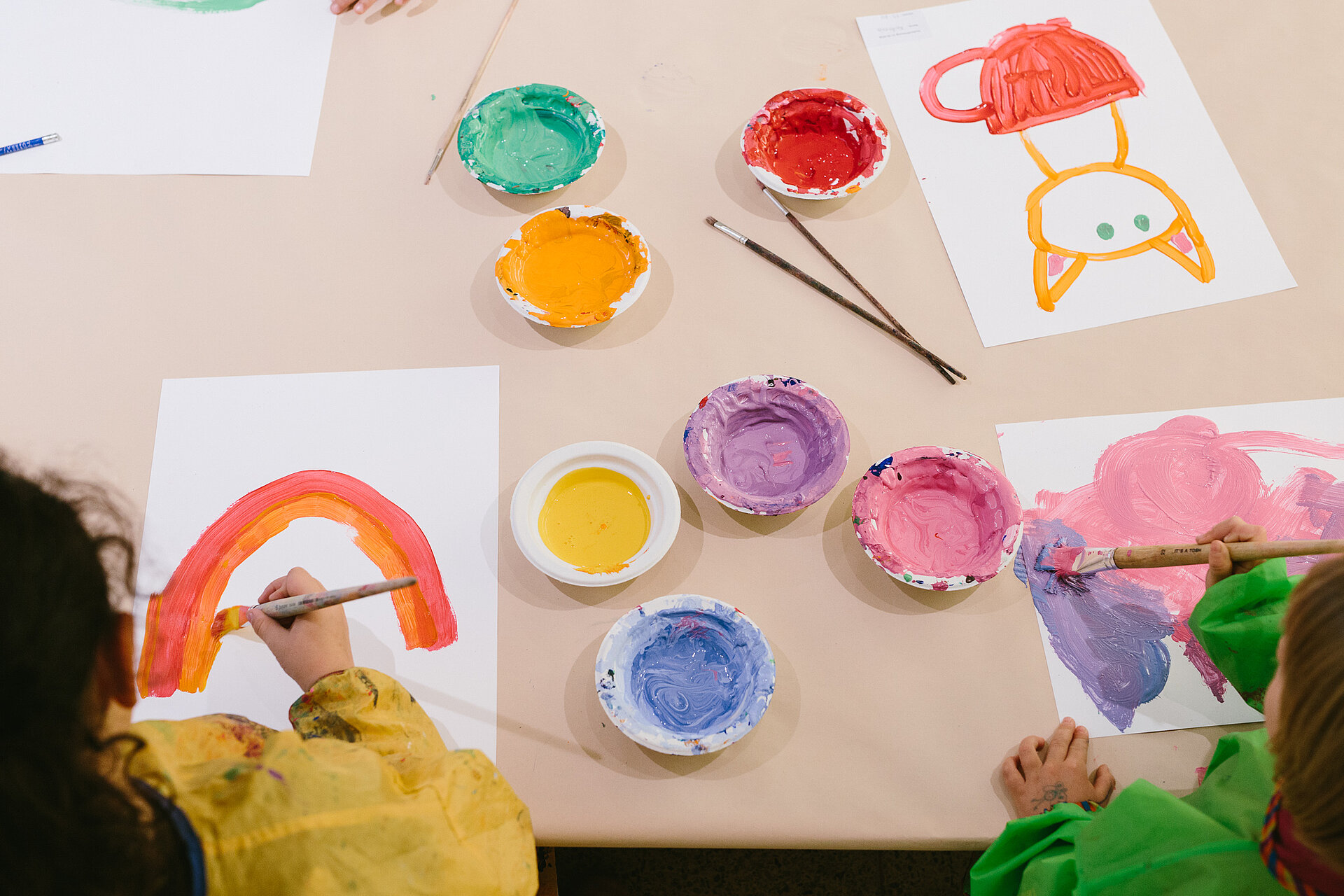 zwei Kinder in gelben und grünen Kitteln sitzen an einem Tisch mit Malutensilien und Farbtöpfen und malen bunte Bilder von Katzen und Regenbogen