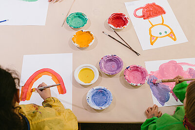 zwei Kinder in gelben und grünen Kitteln sitzen an einem Tisch mit Malutensilien und Farbtöpfen und malen bunte Bilder von Katzen und Regenbogen