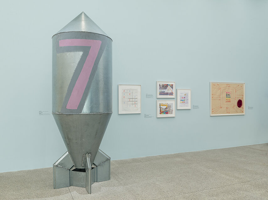 Light blue wall with small pictures on the right, sculpture of a rocket with the number 7 painted in pink on the left in the foreground