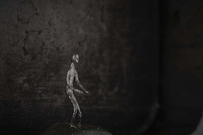 Kleine metallische, menschliche Figur steht auf einer dunklen Oberfläche vor unscharfem, dunklem Hintergrund. / Small metallic human figure standing on a dark surface against a softly blurred dark background.