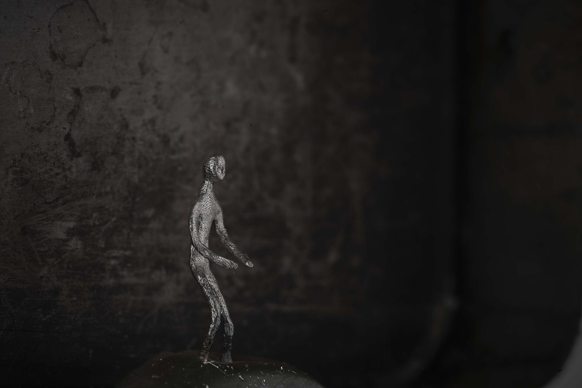 Kleine metallische, menschliche Figur steht auf einer dunklen Oberfläche vor unscharfem, dunklem Hintergrund. / Small metallic human figure standing on a dark surface against a softly blurred dark background.
