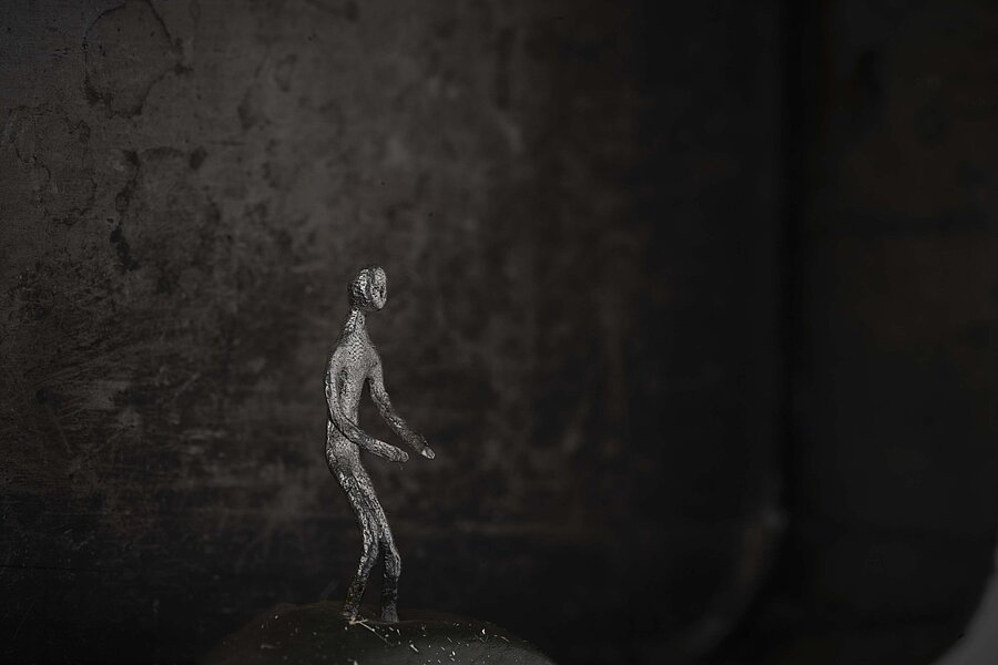 Kleine metallische, menschliche Figur steht auf einer dunklen Oberfläche vor unscharfem, dunklem Hintergrund. / Small metallic human figure standing on a dark surface against a softly blurred dark background.